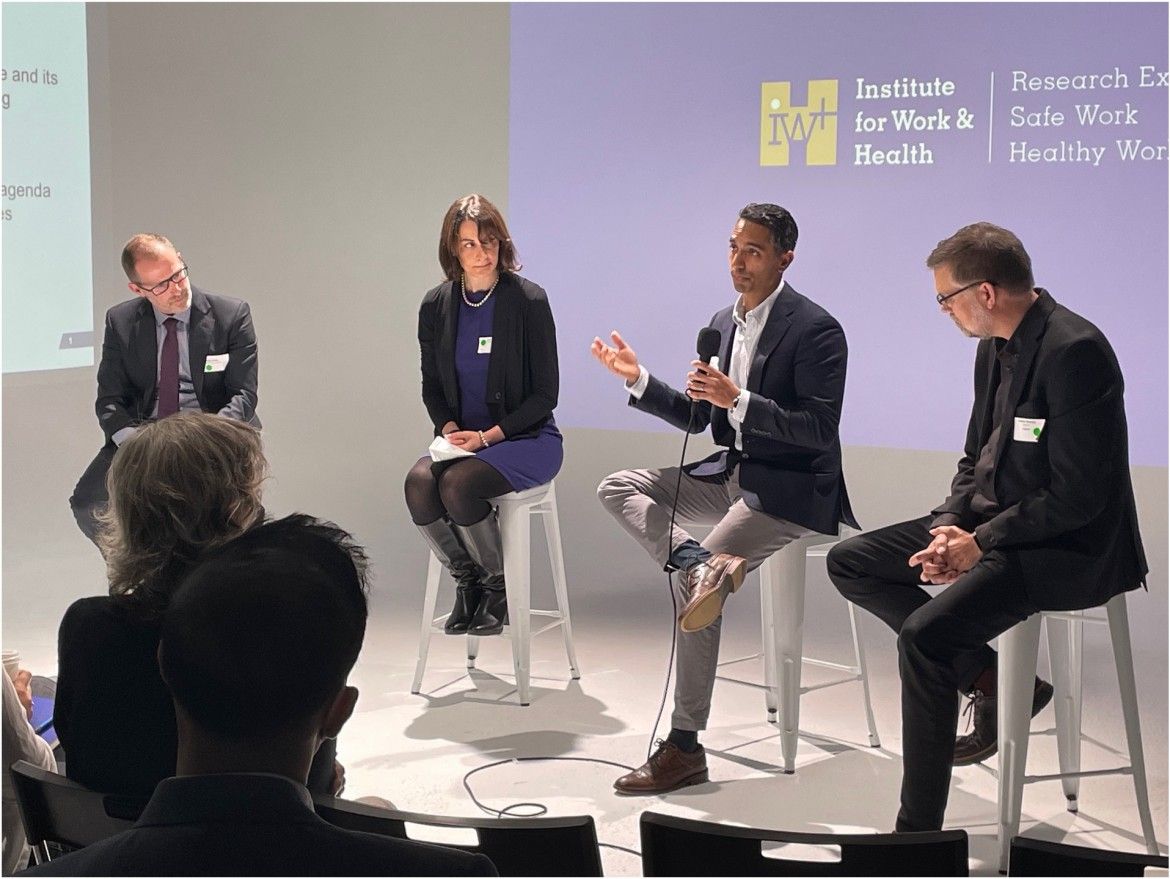 Four people are in discussion as they sit on stools, facing an audience, in front of a screen showing the IWH logo