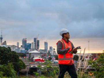 A New Zealand construction worker holding papers looking off-camera with a city skyline behind