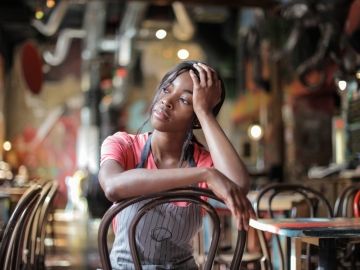 A woman worker in an apron sits inside a restaurant with her head in her hand.