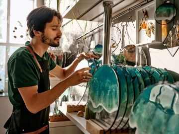 A bearded male worker in handles decorative blue-teal glass displayed on a shelf