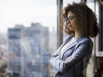 A woman dressed in work attire looks out a window with her arms folded.