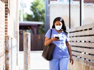 A healthcare worker in scrubs and a mask walks to work with her bag and phone in-hand.