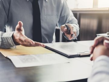 Close up of two pairs individuals sitting across each other at an office desk, one extending a hand to the other