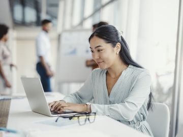 A woman sits in a meeting room at a table working on her laptop computer. Her coworkers are in the background.