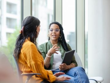 A seated woman with a clipboard in-hand speaks to a female client