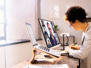 A woman takes notes at a desk while attending a videoconference on the computer monitor