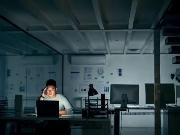 A man works at a computer in an dark, empty office late at night, with his head in his hand