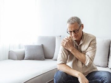 A man sitting on a couch holds his shoulder in pain