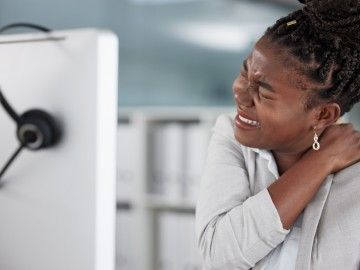 A young worker at her computer workstation holds her shoulder and neck in pain