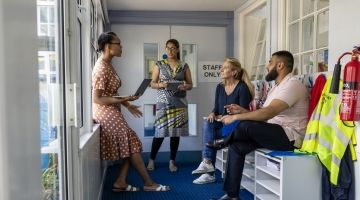 A group of teachers meet outside of a staff room. One holds a clipboard.