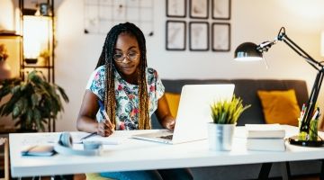 A woman sits at her desk with a laptop and takes notes.