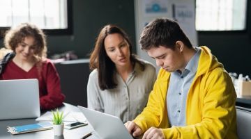 A young man working on a laptop as a female instructor guides him