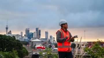 A New Zealand construction worker holding papers looking off-camera with a city skyline behind