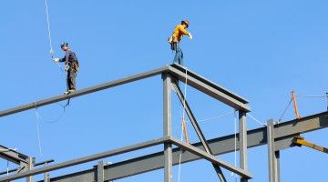 Two ironworkers walking on steel beams against a blue sky