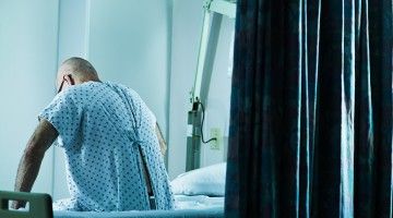 An elderly man in a hospital gown sits on a hospital bed, facing away