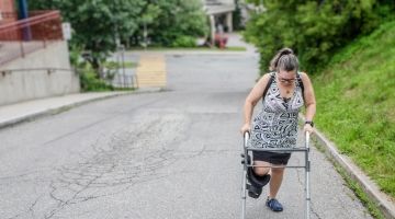 A woman with a leg cast and walker walks up a road.