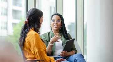 A seated woman with a clipboard in-hand speaks to a female client