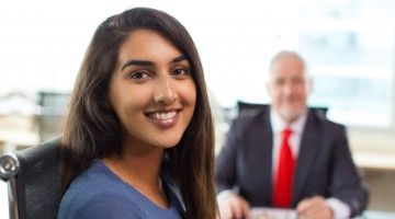 A young woman sits in front of the desk of an older man in a suit and tie, both smiling