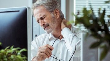 A businessman sits in his office in front of his computer, rubbing his neck in pain