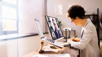 A woman takes notes at a desk while attending a videoconference on the computer monitor