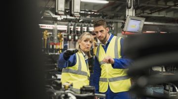 Two workers in safety vests, in a factory environment, point to something offscreen