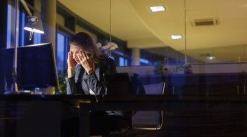 A tired worker holds her head in her hands as she sits at her desk in a dark office