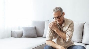 A man sitting on a couch holds his shoulder in pain