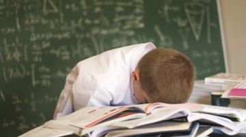 A boy has his head down on his math books, in a despairing manner, with complicated math equations on the blackboard behind him