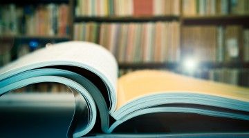 Three open journals lie on a desk in front of shelves of journals behind