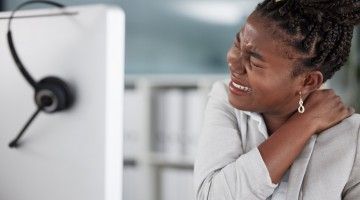 A young worker at her computer workstation holds her shoulder and neck in pain