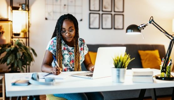 A woman sits at her desk with a laptop and takes notes.