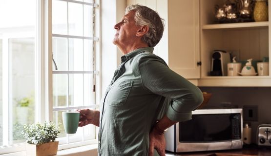 A man stands in his home with a cup of tea while holding his back in pain.