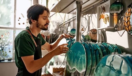 A bearded male worker in an apron handles decorative blue-teal glass discs displayed on a shelf