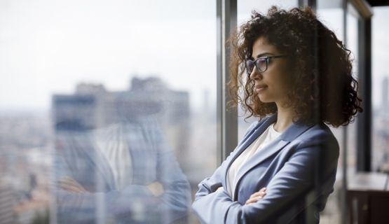 A woman dressed in work attire looks out a window with her arms folded.