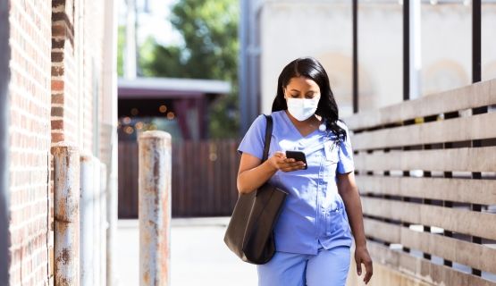 A healthcare worker in scrubs and a mask walks to work with her bag and phone in-hand.