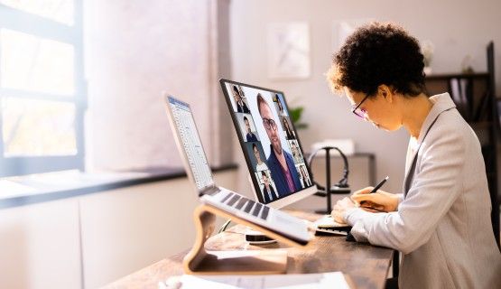 A woman takes notes at a desk while attending a videoconference on the computer monitor