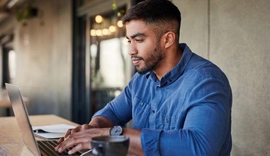 A man in a blue shirt works at a laptop on a long table
