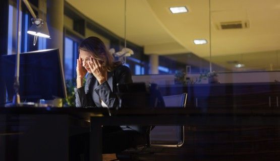 A tired worker holds her head in her hands as she sits at her desk in a dark office