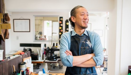 Japanese craftsperson stands in front of his workstation, smiling