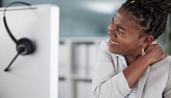 A young worker at her computer workstation holds her shoulder and neck in pain