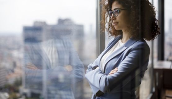 A woman dressed in work attire looks out a window with her arms folded.