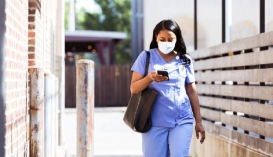A healthcare worker in scrubs and a mask walks to work with her bag and phone in-hand.