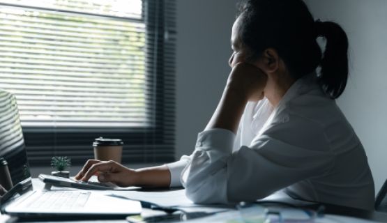 A woman working at a laptop looks out the window with her head resting on her hand