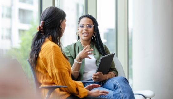 A seated woman with a clipboard in-hand speaks to a female client