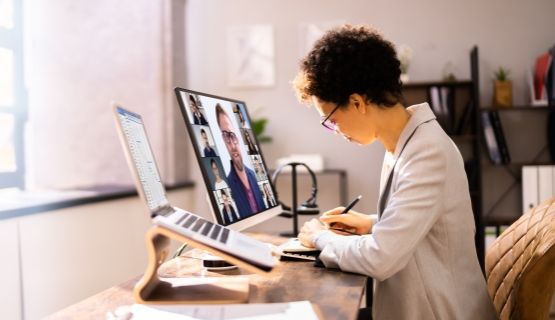 A woman takes notes at a desk while attending a videoconference on the computer monitor