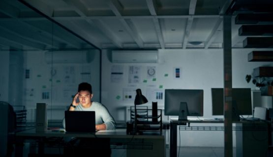 A man works at a computer in an dark, empty office late at night, with his head in his hand 