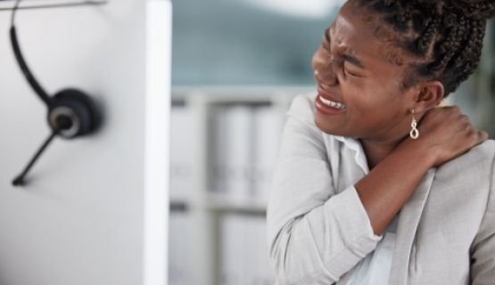 A young worker at her computer workstation holds her shoulder and neck in pain